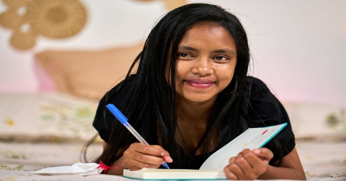 A woman sitting on a bed in a cozy bedroom at night, journaling and smiling.