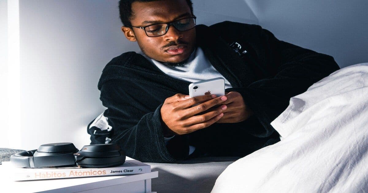 A young man lying on his bed at night, scrolling on his smartphone under warm, dim lighting
