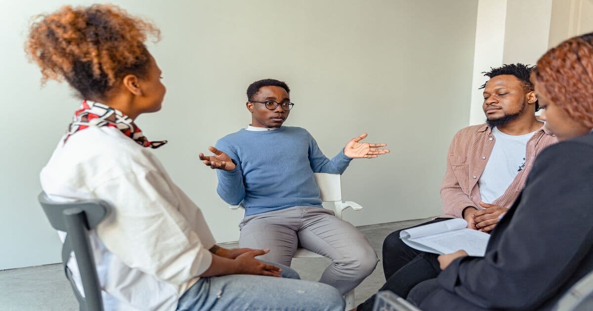 People sitting in a circle as one man speaks with open hands while others listen.