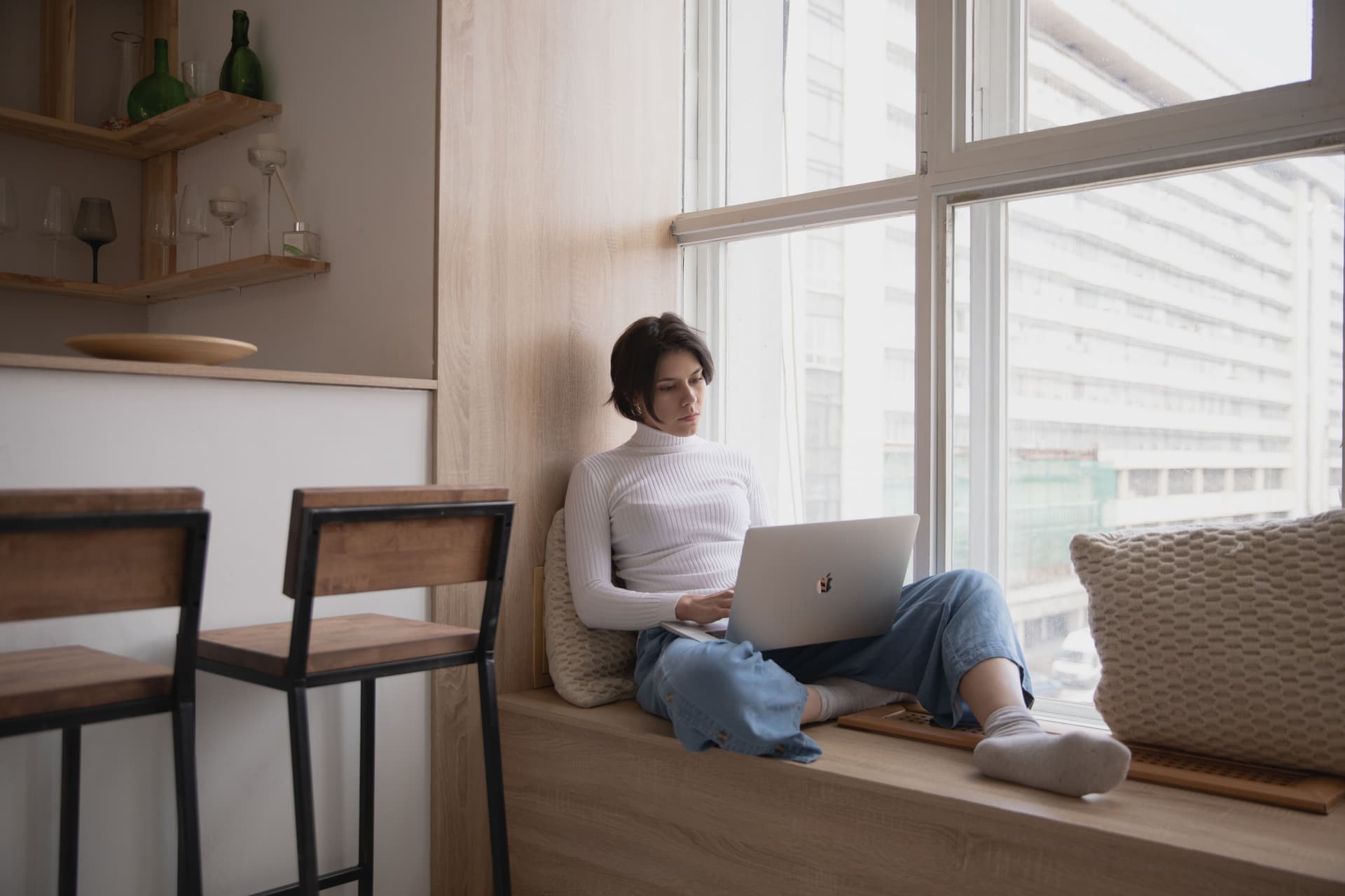 Woman doing remote work on a laptop from a sunlit window seat at home